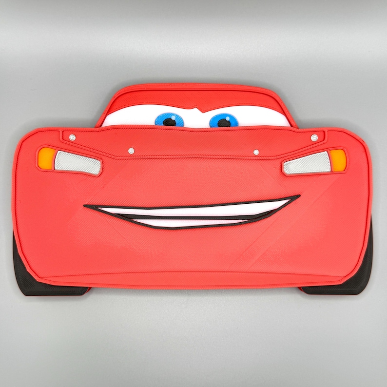 May include: A red, car-shaped flat object featuring a wide, smiling mouth, large white eyes with blue pupils, and yellow and silver headlight details. The design includes black tire accents at the bottom corners and a smooth, molded surface.