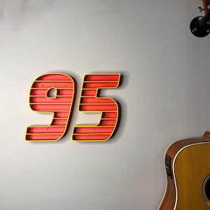 May include: A red and yellow number 95 wall decoration. The numbers are outlined in yellow and have a red interior. The numbers are mounted on a gray wall. A guitar is visible in the background.
