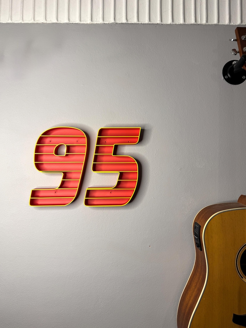 May include: A red and yellow number 95 wall decoration. The numbers are outlined in yellow and have a red interior. The numbers are mounted on a gray wall. A guitar is visible in the background.
