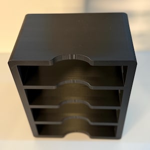 May include: A black, four-tiered desk organizer with curved cutouts on the front of each tier. The organizer is made of a solid, matte material and is designed to hold papers or small items. The overall shape is a cube.