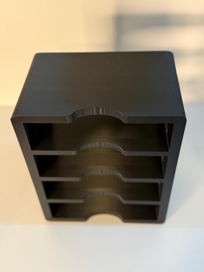 May include: A black, four-tiered desk organizer with curved cutouts on the front of each tier. The organizer is made of a solid, matte material and is designed to hold papers or small items. The overall shape is a cube.