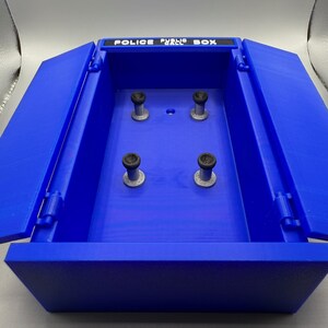 May include: A blue Police Public Box with the top open, revealing the interior. The box has four cylindrical posts inside. The text "POLICE PUBLIC BOX" is printed on the top of the box.