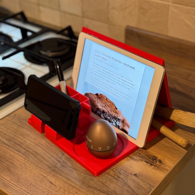 red 3D-printed kitchen organiser used as a recipe book and tablet stand, holding an iPad displaying a recipe. Includes phone holder, pen storage, and a built-in cup holder. Ideal for hands-free cooking and decluttering worktops.