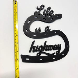 May include: Black decorative wall art in the shape of a horseshoe with the words "Life is a highway". The art measures approximately 10 inches in length. The art is made of a solid material and is suitable for home decor.