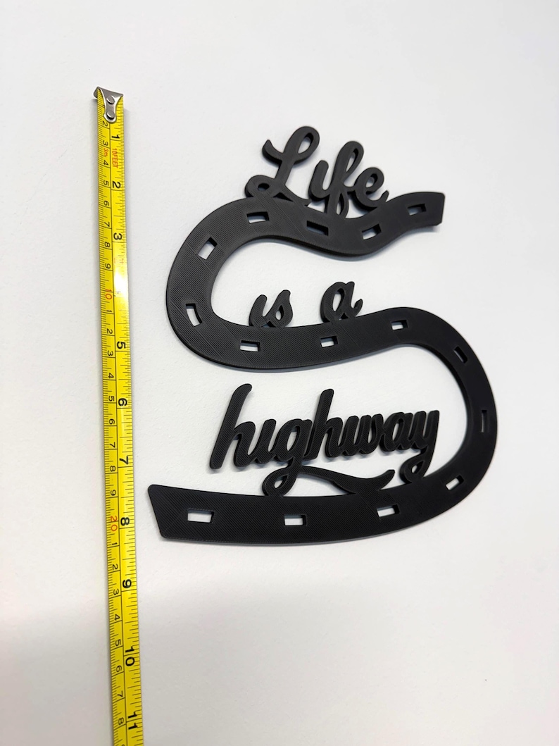 May include: Black decorative wall art in the shape of a horseshoe with the words "Life is a highway". The art measures approximately 10 inches in length. The art is made of a solid material and is suitable for home decor.