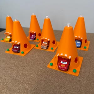 May include: A collection of orange traffic cone-shaped toy structures, each with a small car inside. The cars are various colors, including red, yellow, purple, and blue. The structures have a white top and are set on a square base.