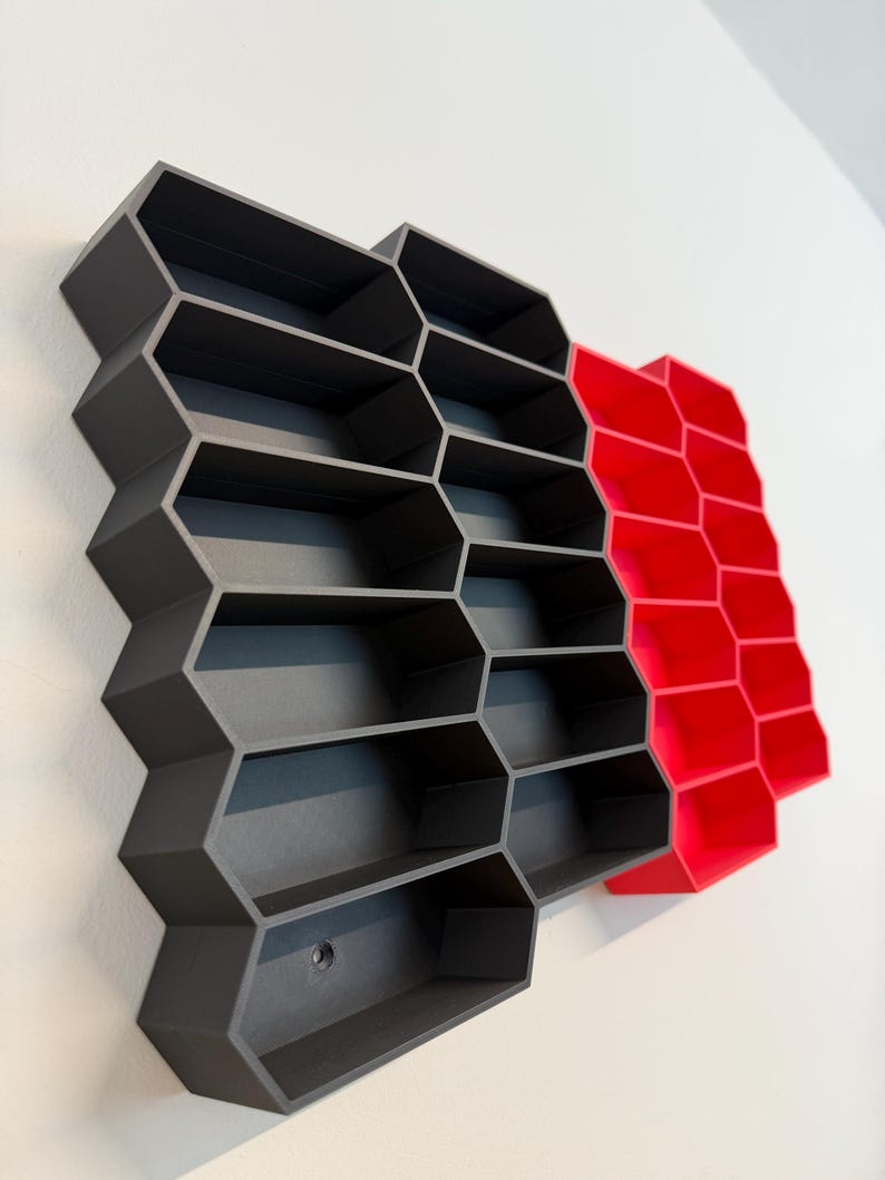 May include: A wall-mounted storage unit featuring a series of interconnected hexagonal compartments. The majority of the structure is matte dark gray, while a smaller section on the right side is a vibrant, solid red. The unit has an open-front design.