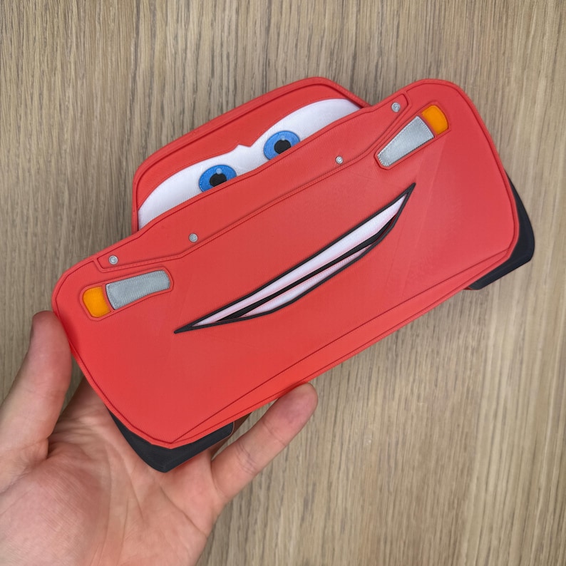 K&ouml;nnte beinhalten: Rotes Lightning McQueen-Auto-Accessoire mit einem l&auml;chelnden Gesicht, blauen Augen und wei&szlig;en Z&auml;hnen. Das Zubeh&ouml;r hat schwarze Akzente und wird in einer Hand gehalten. Das Auto-Design umfasst Scheinwerfer und eine Windschutzscheibe.