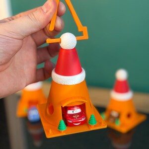 May include: Orange traffic cone ornament with a red Santa hat and a small red car inside. The ornament has a white pom-pom and a yellow handle. Two small green trees flank the car. Additional similar ornaments are in the background.