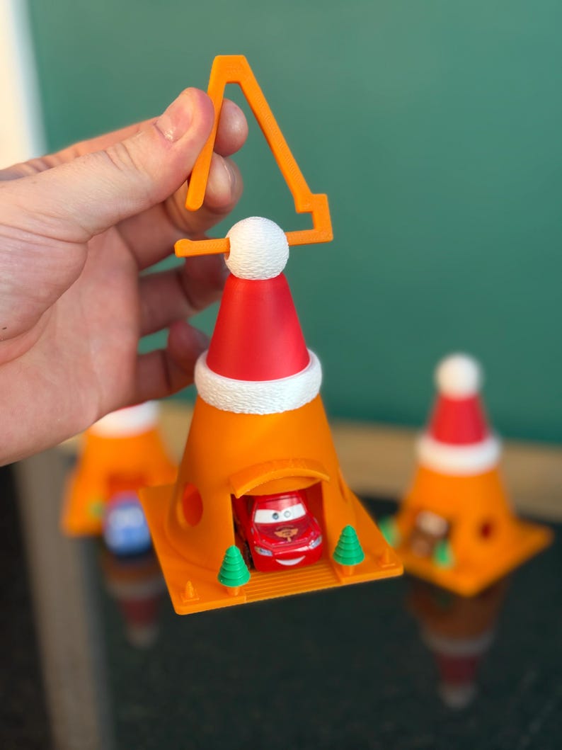 May include: Orange traffic cone ornament with a red Santa hat and a small red car inside. The ornament has a white pom-pom and a yellow handle. Two small green trees flank the car. Additional similar ornaments are in the background.