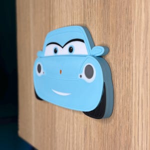 May include: A light blue cartoon car-shaped decoration with a smiling face, black tires, and white accents. The car is affixed to a wooden surface, creating a playful and decorative element.