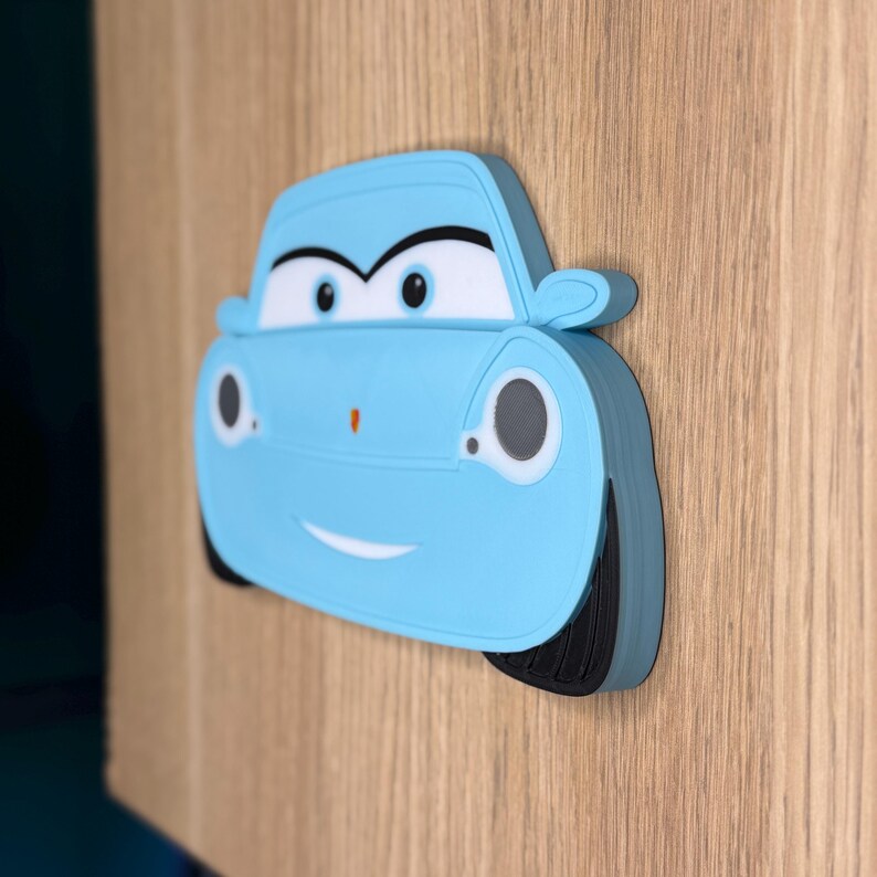 May include: A light blue cartoon car-shaped decoration with a smiling face, black tires, and white accents. The car is affixed to a wooden surface, creating a playful and decorative element.