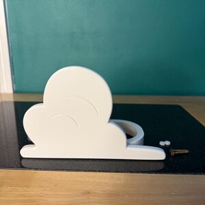 May include: A white cloud-shaped shelf with curved details, likely for decorative purposes. Includes small screws for mounting. The shelf is on a black surface with a teal background.