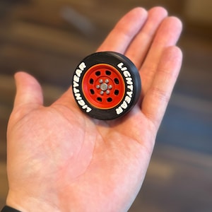 May include: A miniature black and red toy car wheel with the word "LIGHTYEAR" in white lettering. The wheel has a red rim and a silver center. The toy is held in a person's hand.