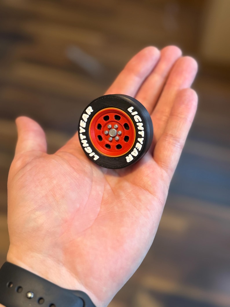 May include: A miniature black and red toy car wheel with the word "LIGHTYEAR" in white lettering. The wheel has a red rim and a silver center. The toy is held in a person's hand.