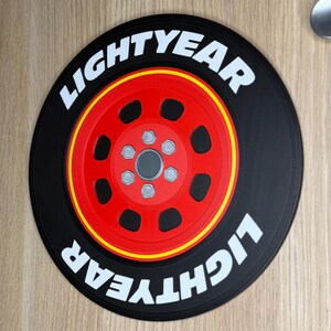 May include: A circular black decorative item featuring a red wheel with a yellow stripe and the text "LIGHTYEAR" in white. The wheel has six silver-colored bolts. The item is likely a themed decoration.