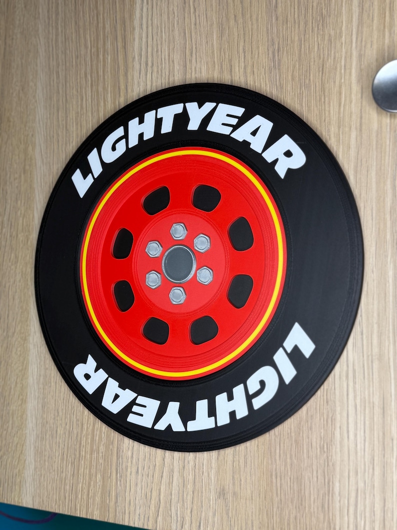 May include: A circular black decorative item featuring a red wheel with a yellow stripe and the text "LIGHTYEAR" in white. The wheel has six silver-colored bolts. The item is likely a themed decoration.