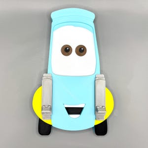 May include: A light blue cartoon car-shaped object with brown eyes, a white face, and a smiling black mouth. The car has a yellow circle on the bottom with black tires and silver side panels. The object is a fun, decorative item.