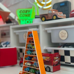 Op de afbeelding: Een oranje, driehoekige displayplank gevuld met miniatuur speelgoedauto's. Op de plank staat de tekst "COZY CONE MOTEL". De auto's zijn in verschillende kleuren en ontwerpen. Op de achtergrond staat een neonbord met de tekst "COZY CONE MOTEL".