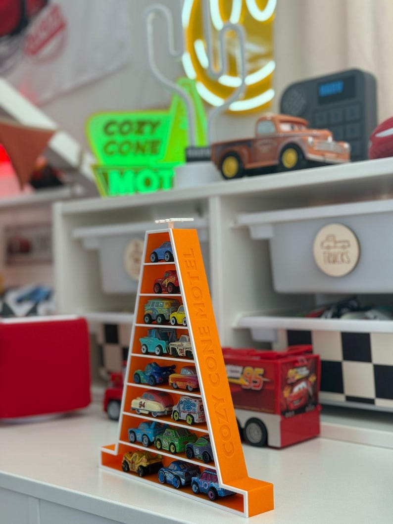 Op de afbeelding: Een oranje, driehoekige displayplank gevuld met miniatuur speelgoedauto's. Op de plank staat de tekst "COZY CONE MOTEL". De auto's zijn in verschillende kleuren en ontwerpen. Op de achtergrond staat een neonbord met de tekst "COZY CONE MOTEL".