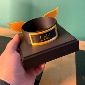 May include: A dark brown circular display stand with a square base, featuring two bright yellow wings attached to the sides. A rectangular yellow frame is mounted on the front of the circular ring, containing the name Loki in cursive script.