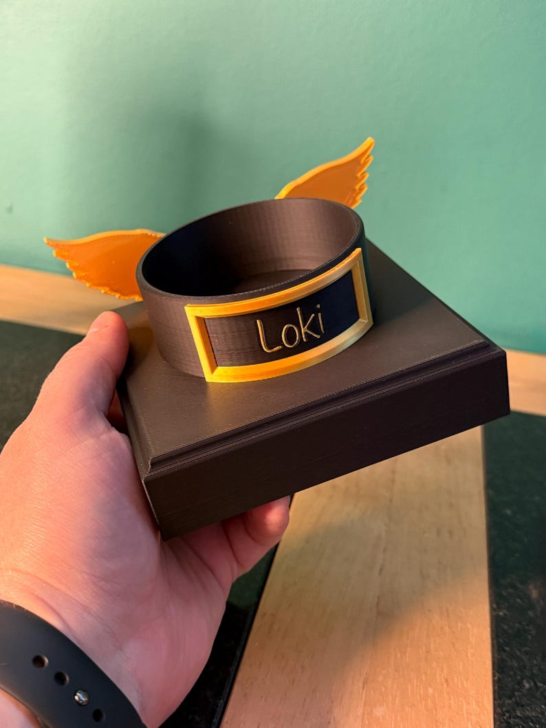 May include: A dark brown circular display stand with a square base, featuring two bright yellow wings attached to the sides. A rectangular yellow frame is mounted on the front of the circular ring, containing the name Loki in cursive script.