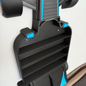 Op de afbeelding: Een zwart en blauw Formule 1-auto-vormig rek met vier planken. Het rek is aan een witte muur bevestigd. De auto heeft zwarte wielen en blauwe accenten. Het rek is ontworpen om kleine items in op te bergen.