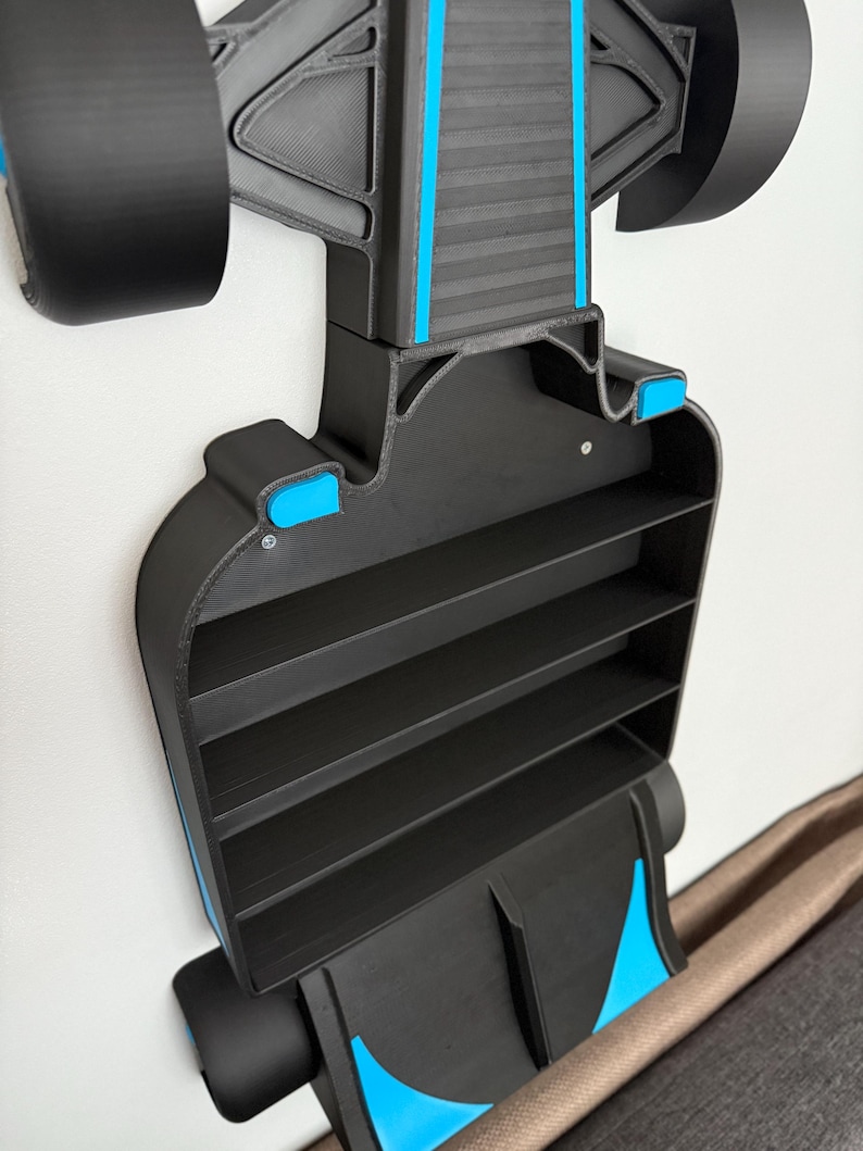 Op de afbeelding: Een zwart en blauw Formule 1-auto-vormig rek met vier planken. Het rek is aan een witte muur bevestigd. De auto heeft zwarte wielen en blauwe accenten. Het rek is ontworpen om kleine items in op te bergen.