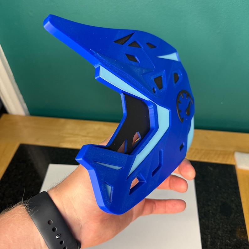 May include: A blue and light blue helmet with a visor, designed for sports. The helmet has a black interior and a circular emblem on the side. The helmet is held in a person's hand.