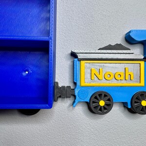 Close-up of a 3D-printed blue and yellow train engine labelled “Noah,” placed next to to a blue toy car shelf. Designed to resemble a train carriage coupling.