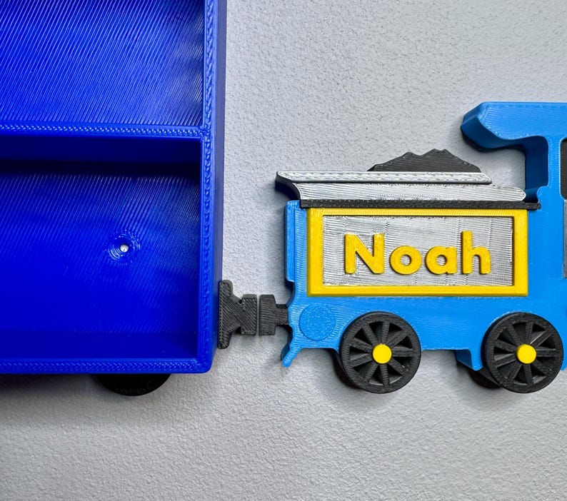 Close-up of a 3D-printed blue and yellow train engine labelled “Noah,” placed next to to a blue toy car shelf. Designed to resemble a train carriage coupling.