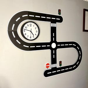 May include: A black and white road-themed wall clock with a circular clock face. The clock is integrated into a road design with a stop sign and traffic lights. The road design is mounted on a gray wall.
