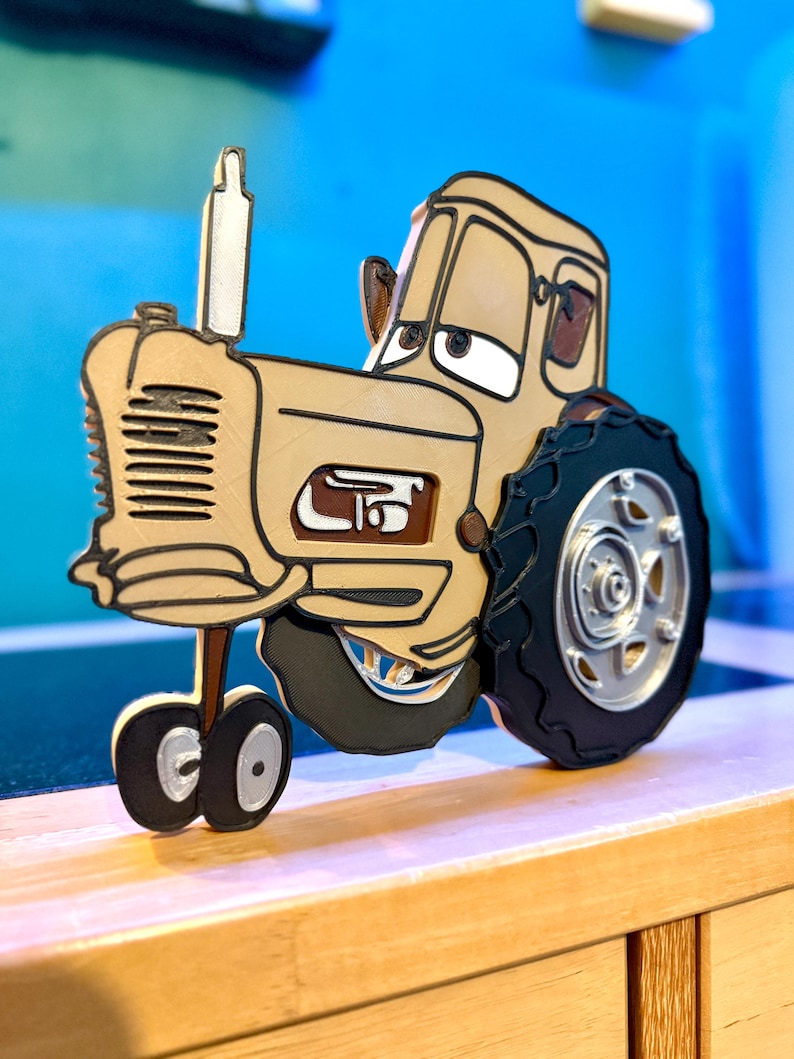 May include: A three-dimensional layered wall art piece depicting a tan tractor with large black tires, a silver exhaust pipe, and expressive eyes on the windshield. The tractor features a detailed front grille, side engine panel, and wheels.