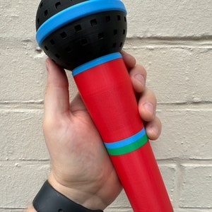 A 3D-printed, colorful plastic microphone prop held in a hand. This vibrant and durable design is perfect for photo booth props, party accessories, or as a fun kids' toy, available in various color combinations.