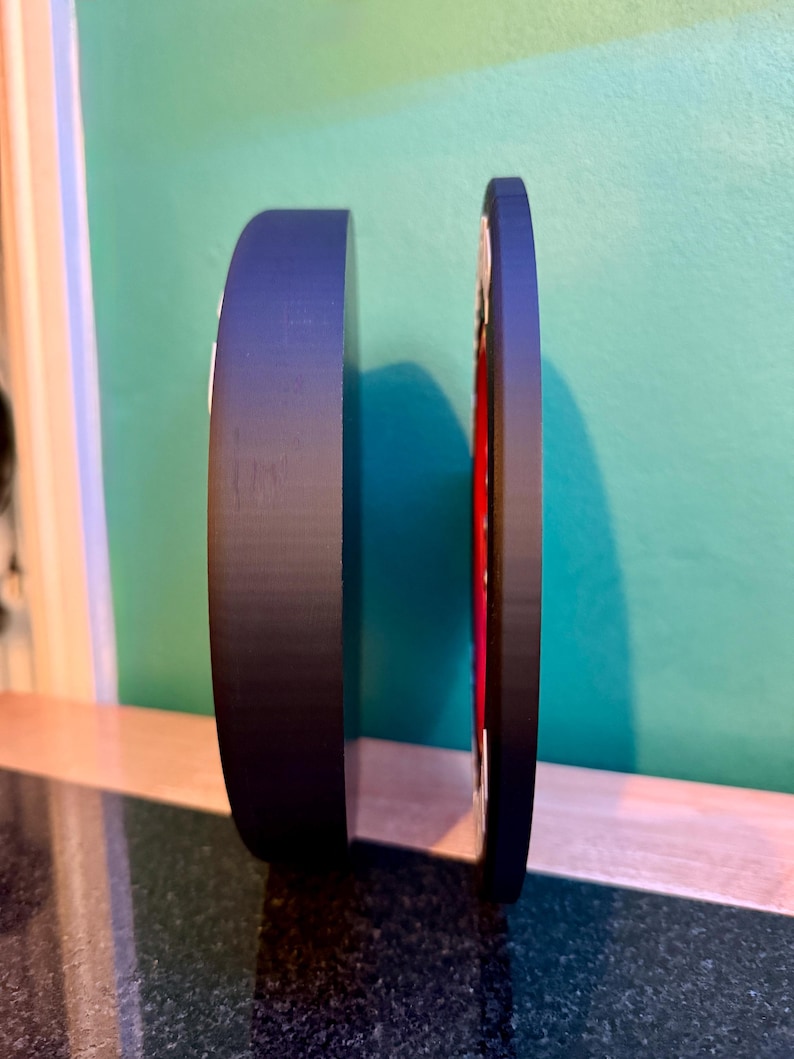 May include: A black, circular object with a red and white design on the side. The object is standing upright on a dark surface, with a green and blue background.