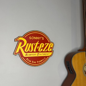 Puede incluir: Un letrero redondo rojo y amarillo con el texto "Sonny's Rust-eze Bumper Ointment Rear End Formula". El letrero est&aacute; montado en una pared gris, con una parte de una guitarra ac&uacute;stica de madera a la derecha.