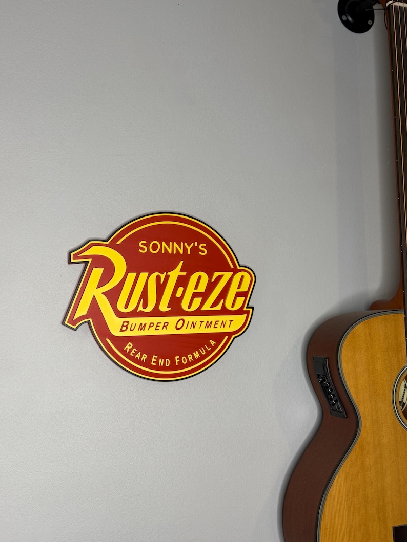 Puede incluir: Un letrero redondo rojo y amarillo con el texto "Sonny's Rust-eze Bumper Ointment Rear End Formula". El letrero est&aacute; montado en una pared gris, con una parte de una guitarra ac&uacute;stica de madera a la derecha.
