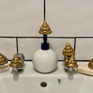 Five gold glittery 3D-printed poop emojis arranged around a white bathroom sink, sitting on the taps, soap, and brush. A playful and eco-friendly novelty decoration made from recycled filament waste.