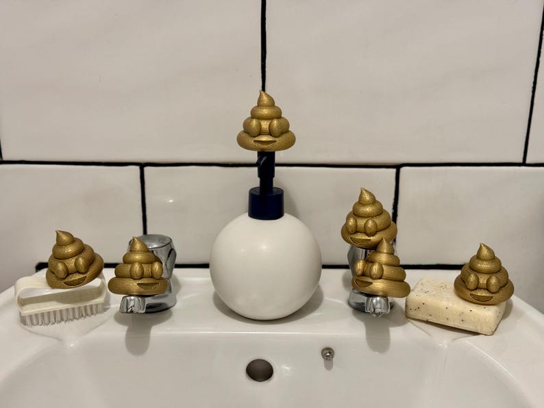 Five gold glittery 3D-printed poop emojis arranged around a white bathroom sink, sitting on the taps, soap, and brush. A playful and eco-friendly novelty decoration made from recycled filament waste.