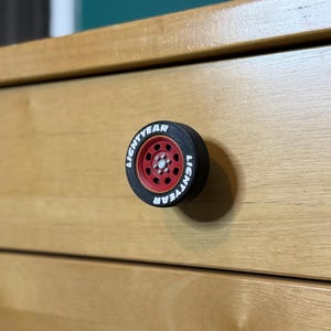 May include: A black and red drawer pull shaped like a tire, with the word "LIGHTYEAR" in white lettering around the rim. The drawer pull is mounted on a light brown wooden drawer.