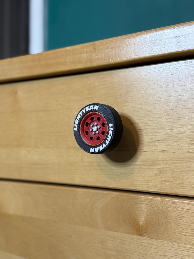 May include: A black and red drawer pull shaped like a tire, with the word "LIGHTYEAR" in white lettering around the rim. The drawer pull is mounted on a light brown wooden drawer.