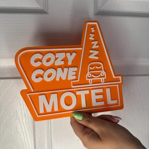 以下が含まれることがあります： オレンジ色のモーテルの看板に、白い文字で「Cozy Cone Motel」と書かれています。看板には、眠そうな顔をしたスタイリッシュな車が描かれており、側面には「zzz」と書かれています。