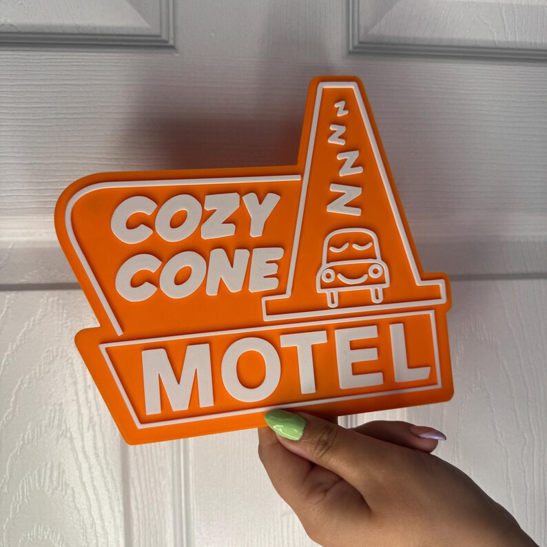 以下が含まれることがあります： オレンジ色のモーテルの看板に、白い文字で「Cozy Cone Motel」と書かれています。看板には、眠そうな顔をしたスタイリッシュな車が描かれており、側面には「zzz」と書かれています。