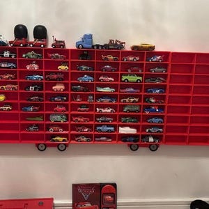 Op de afbeelding: Rode speelgoedtruck-rek met meerdere planken om speelgoedauto's te tonen. De planken zijn gevuld met verschillende rode, blauwe en gele speelgoedauto's. De truck is gelabeld met het nummer "95".
