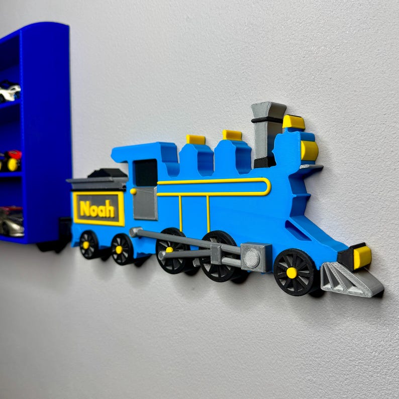 A 3D-printed blue toy train display shelf, mounted on the wall and filled with die-cast cars. The front train engine is labelled with the name “Noah” in yellow, styled like a locomotive pulling the shelf.