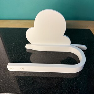 May include: A white, cloud-shaped toilet paper holder. The holder has a curved bar for the toilet paper roll and a cloud-shaped backplate. The holder is made of a solid, smooth material and is designed to be mounted on a wall.
