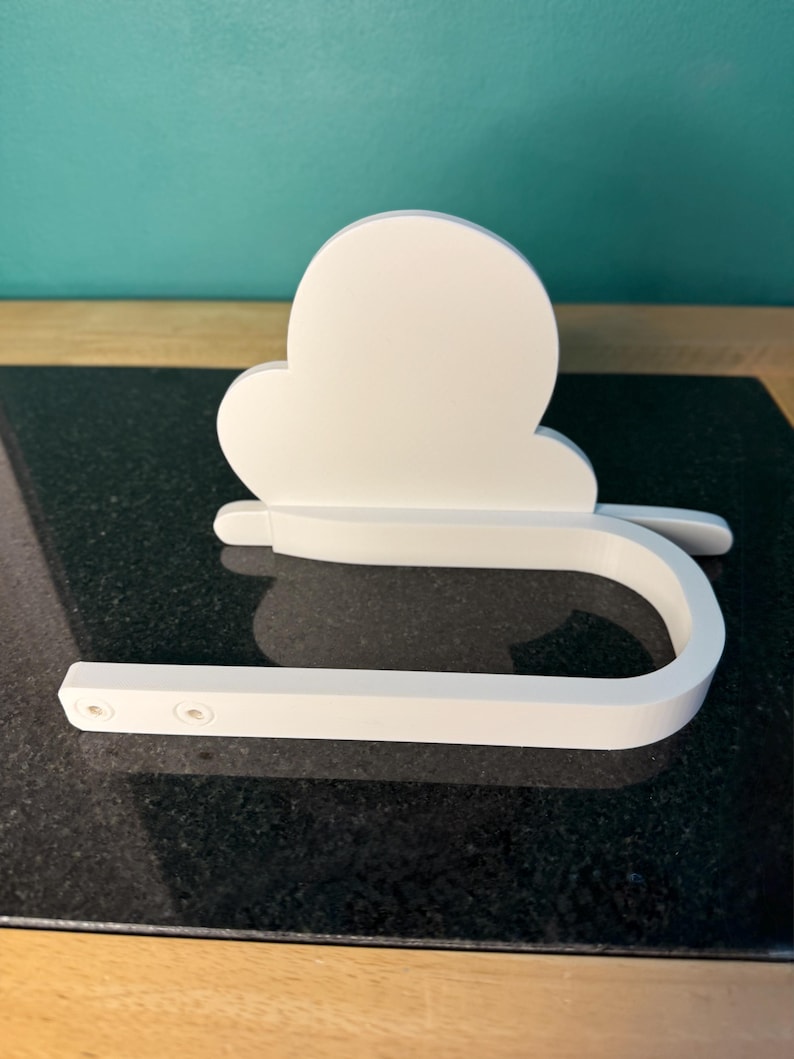 May include: A white, cloud-shaped toilet paper holder. The holder has a curved bar for the toilet paper roll and a cloud-shaped backplate. The holder is made of a solid, smooth material and is designed to be mounted on a wall.