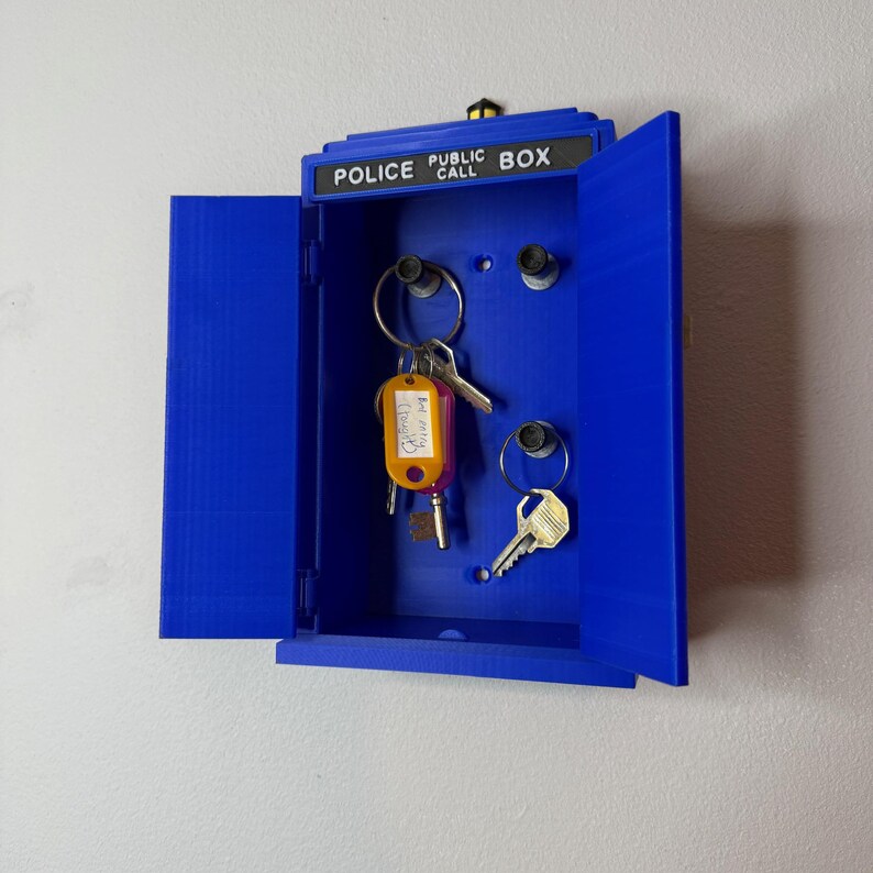 May include: A blue key holder shaped like a police call box, with the words "POLICE CALL BOX" across the top. The key holder is open, revealing keys hanging from metal rings. The keys have various shapes and colors.