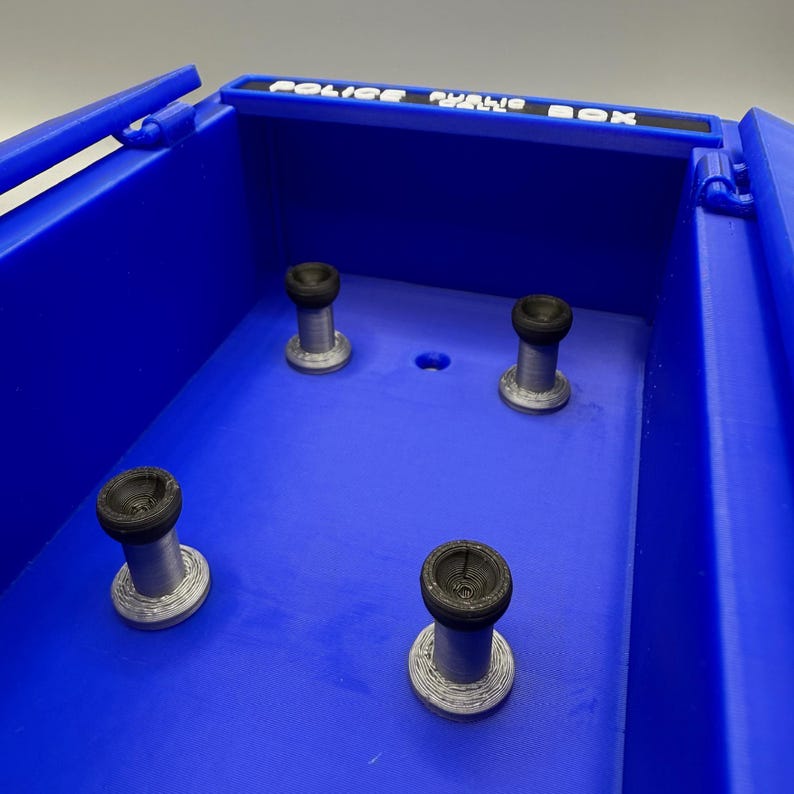 May include: A blue, rectangular box with the words "POLICE PUBLIC CALL BOX" in white lettering across the top. Inside, four cylindrical objects with black tops and silver bases are visible.