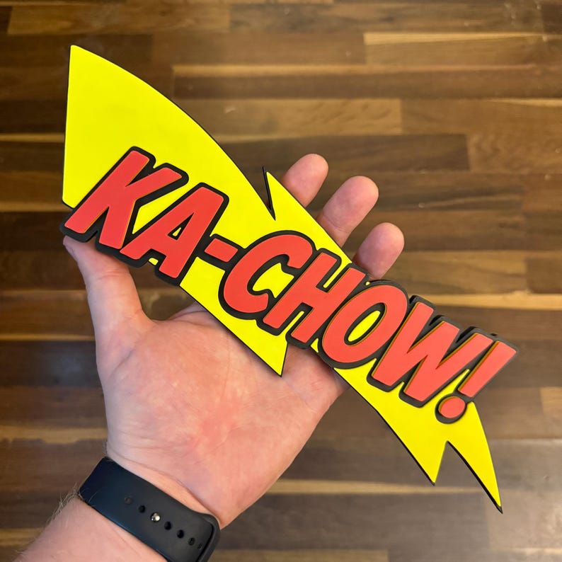 May include: A yellow lightning bolt-shaped sign with the words "KA-CHOW!" in red, outlined in black. The sign is held in a hand, and the background is a wooden surface.